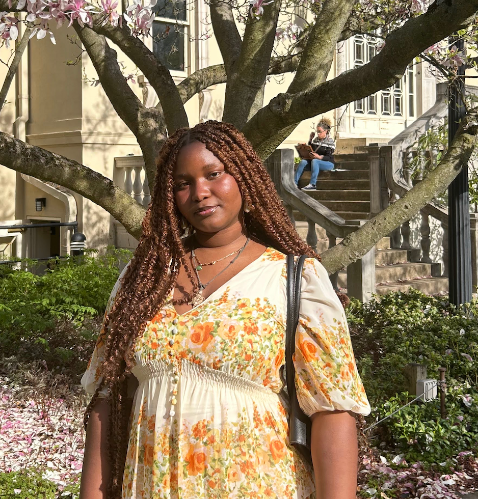 Melissa Munroe poses for a headshot infront of a tree. She is wearing a orange and cream colored dress. 